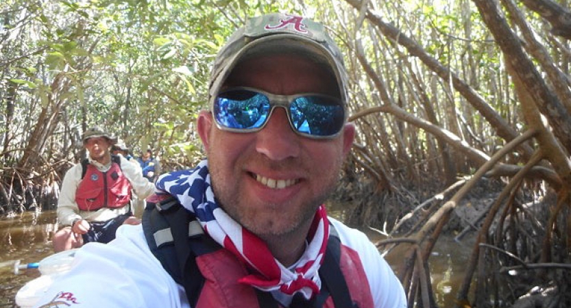 a person in a hat, sunglasses and an american flag bandana smiles from a canoe on an outward bound veterans expedition 